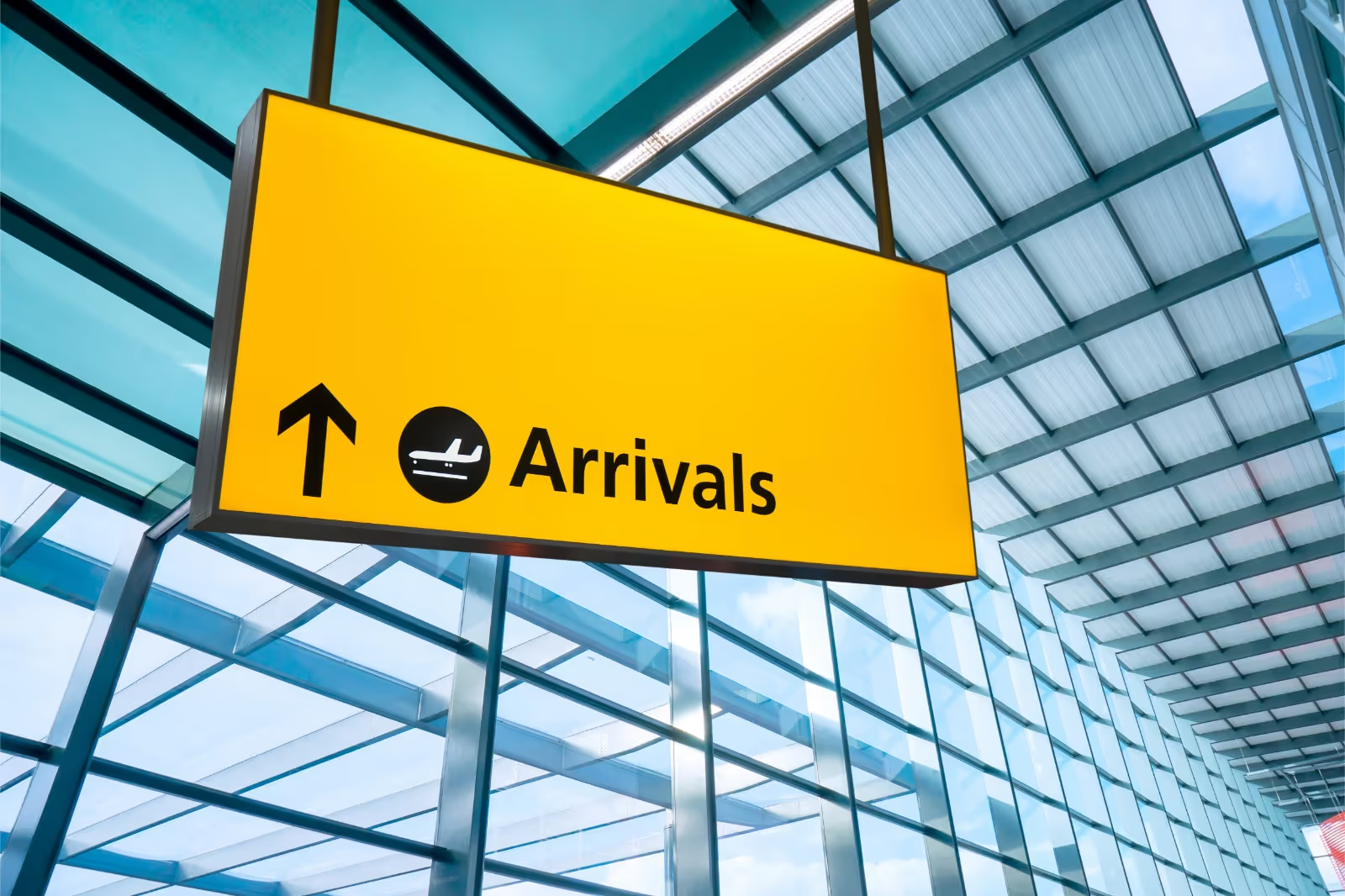 Geel bord met de tekst 'Arrivals', een pijl omhoog en een vliegtuigicoon, hangend in een luchthaventerminal met glazen dak
