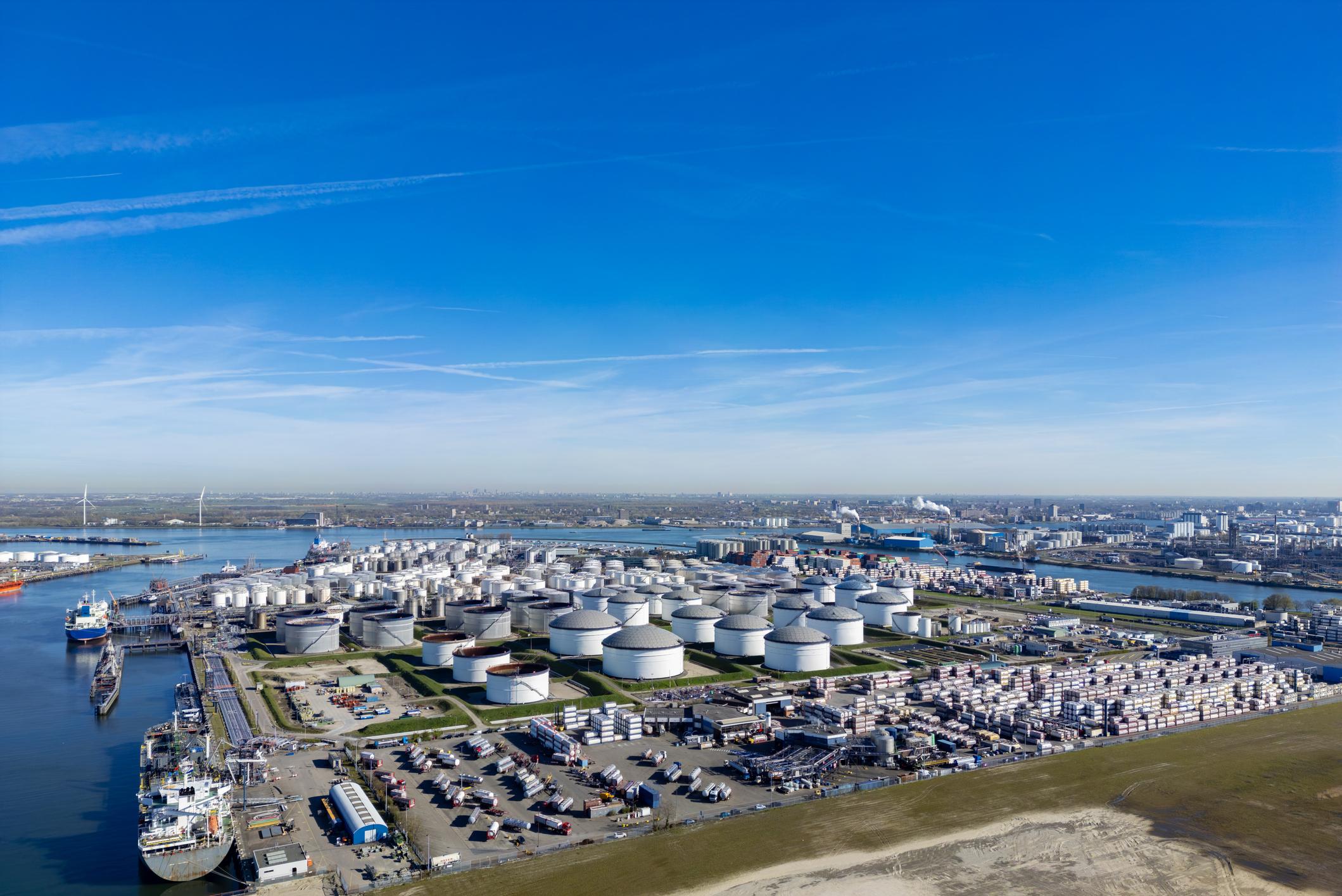 Luchtfoto van een havengebied met opslagtanks, containers en aangemeerde schepen onder een blauwe lucht