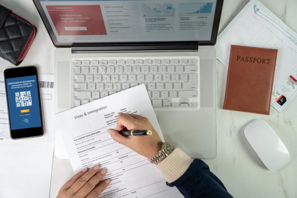 Iemand vult een visum- en immigratieformulier in, met een paspoort, telefoon met vaccinatiecertificaat en laptop op tafel.