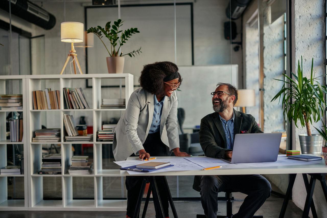 Twee lachende collega's overleggen samen aan een bureau met laptop en documenten in een modern kantoor
