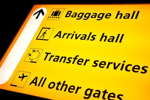 Geel verlicht bewegwijzeringsbord op een luchthaven met pictogrammen en richtingen naar Baggage hall, Arrivals hall, Transfer services en All other gates