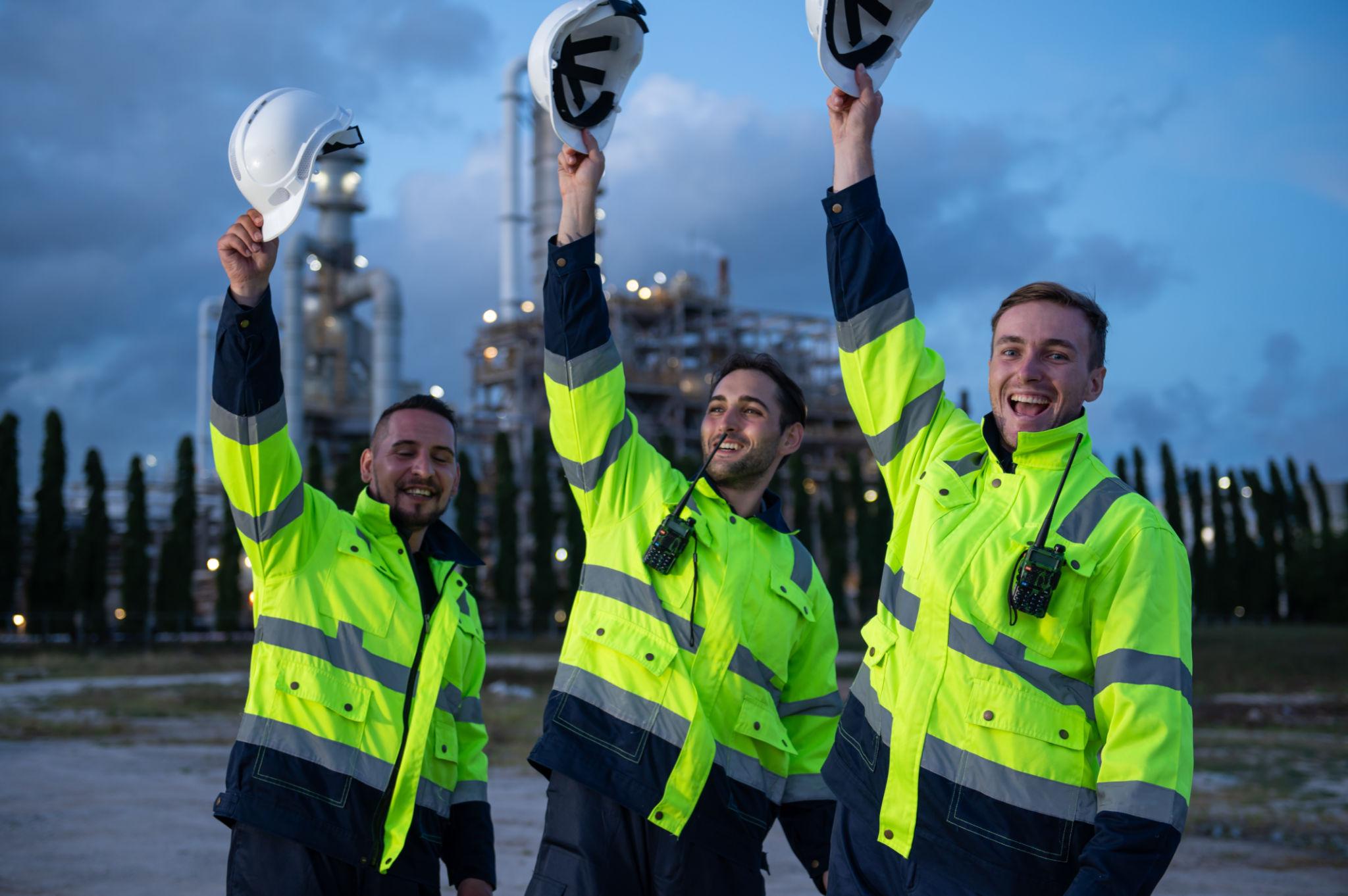 Drie juichende werknemers in veiligheidskleding houden hun witte helmen omhoog bij een industriële installatie